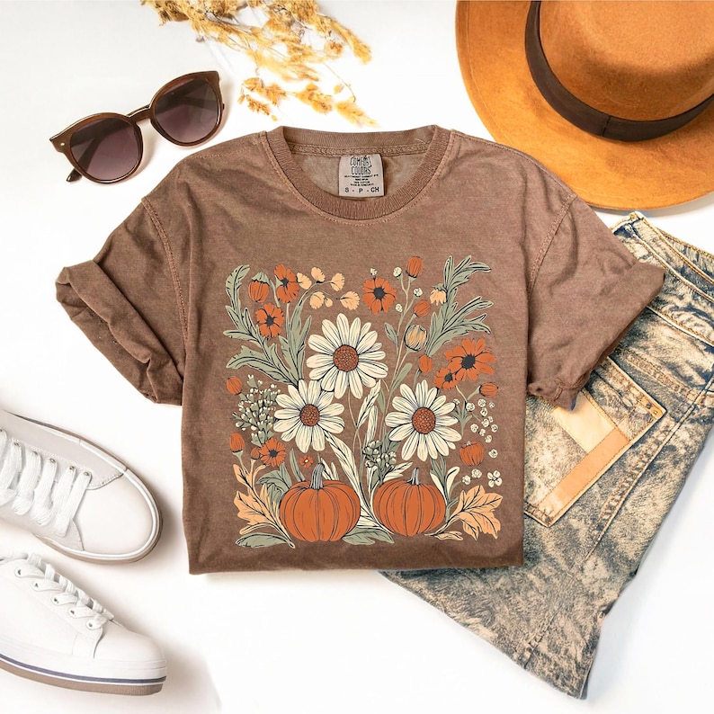 May include: Brown t-shirt with a floral and pumpkin design. The graphic features large white daisies, orange flowers, and two orange pumpkins. The shirt is paired with sunglasses, a hat, and jeans.