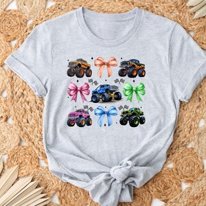 Puede incluir: Una camiseta gris claro con un gr&aacute;fico colorido de monster trucks y lazos. El dise&ntilde;o incluye seis monster trucks en varios colores, como naranja, azul y verde, combinados con lazos a juego. La camiseta se muestra sobre una alfombra tejida.