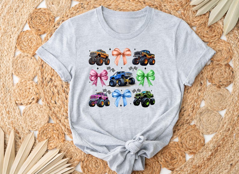 Puede incluir: Una camiseta gris claro con un gr&aacute;fico colorido de monster trucks y lazos. El dise&ntilde;o incluye seis monster trucks en varios colores, como naranja, azul y verde, combinados con lazos a juego. La camiseta se muestra sobre una alfombra tejida.