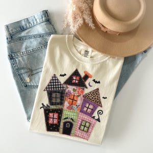 May include: A cream-colored Comfort Colors t-shirt featuring a patchwork design of colorful houses, bats, and a crescent moon. The shirt is paired with light-wash denim jeans, a tan hat, and a gold necklace.