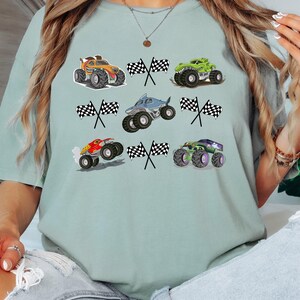 Pode incluir: T-shirt verde claro com ilustra&ccedil;&otilde;es de monster trucks e bandeiras quadriculadas. Os caminh&otilde;es s&atilde;o nas cores laranja, verde, vermelho e roxo. O design &eacute; divertido e adequado para crian&ccedil;as.