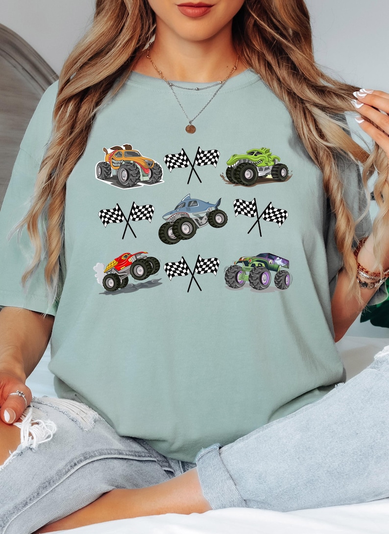Pode incluir: T-shirt verde claro com ilustra&ccedil;&otilde;es de monster trucks e bandeiras quadriculadas. Os caminh&otilde;es s&atilde;o nas cores laranja, verde, vermelho e roxo. O design &eacute; divertido e adequado para crian&ccedil;as.