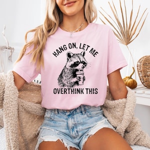 May include: A light pink t-shirt featuring a black and white illustration of a raccoon holding a coffee cup. The text "HANG ON, LET ME OVERTHINK THIS" is printed above and below the raccoon image. The shirt is a short-sleeved crew neck.