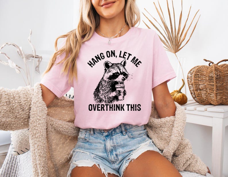 May include: A light pink t-shirt featuring a black and white illustration of a raccoon holding a coffee cup. The text "HANG ON, LET ME OVERTHINK THIS" is printed above and below the raccoon image. The shirt is a short-sleeved crew neck.