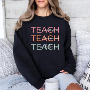 May include: Black sweatshirt with the words "Teach" repeated three times in different colors, with the words "compassion", "kindness", and "confidence" written in smaller font underneath each "Teach".