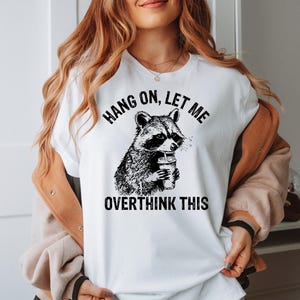 May include: White t-shirt featuring a black and white illustration of a raccoon holding a coffee cup. The text "HANG ON, LET ME OVERTHINK THIS" is printed above and below the raccoon graphic. The shirt has short sleeves.