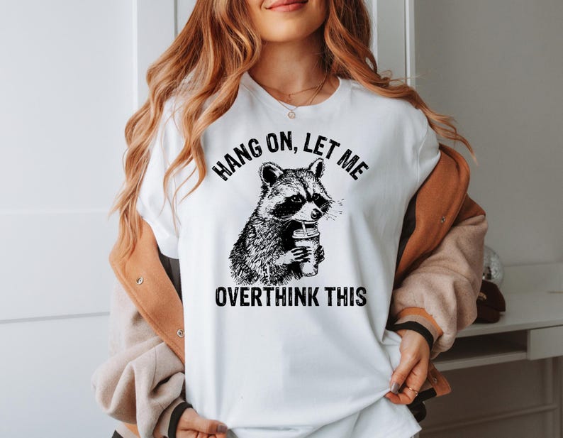 May include: White t-shirt featuring a black and white illustration of a raccoon holding a coffee cup. The text "HANG ON, LET ME OVERTHINK THIS" is printed above and below the raccoon graphic. The shirt has short sleeves.