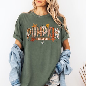 Pu&ograve; includere: T-shirt verde oliva con la scritta "PUMPKIN SEASON" in un design decorativo a tema autunnale. Il testo incorpora motivi a quadri, floreali e a foglie. La maglietta ha le maniche arrotolate ed &egrave; abbinata a una giacca di jeans.