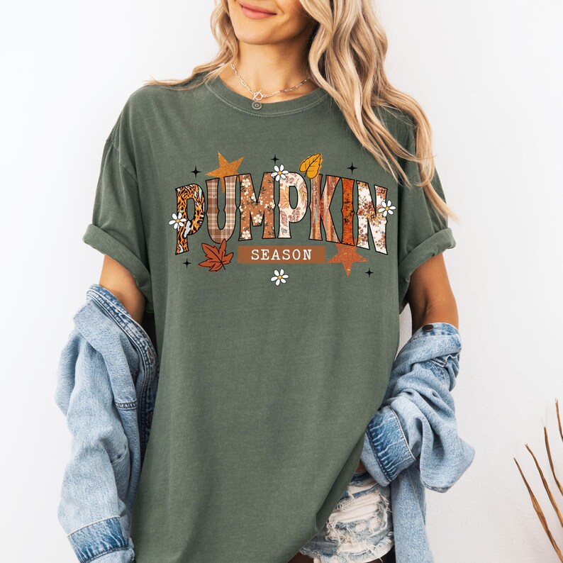 Pu&ograve; includere: T-shirt verde oliva con la scritta "PUMPKIN SEASON" in un design decorativo a tema autunnale. Il testo incorpora motivi a quadri, floreali e a foglie. La maglietta ha le maniche arrotolate ed &egrave; abbinata a una giacca di jeans.