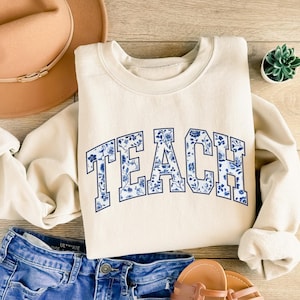 May include: A cream-colored sweatshirt with a blue and white floral design that says "TEACH" in a collegiate font.