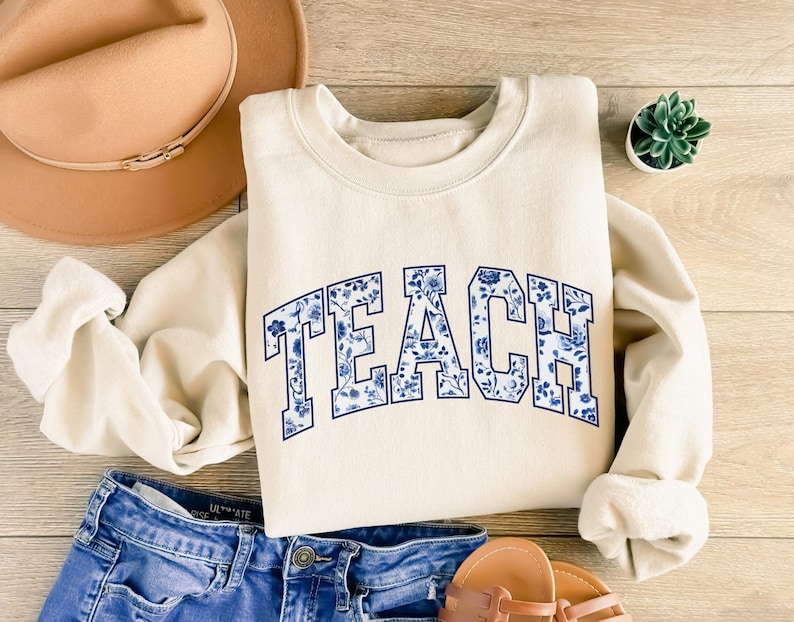 May include: A cream-colored sweatshirt with a blue and white floral design that says "TEACH" in a collegiate font.