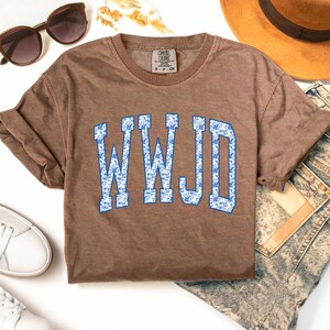May include: A brown short-sleeved t-shirt with the text "WWJD" in blue letters with a floral pattern. The shirt is styled with sunglasses, a hat, white sneakers, and denim jeans.