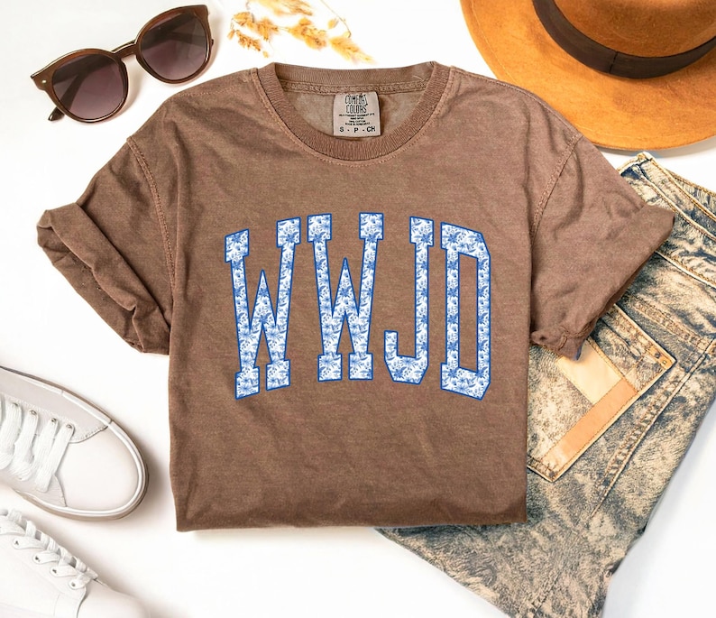 May include: A brown short-sleeved t-shirt with the text "WWJD" in blue letters with a floral pattern. The shirt is styled with sunglasses, a hat, white sneakers, and denim jeans.