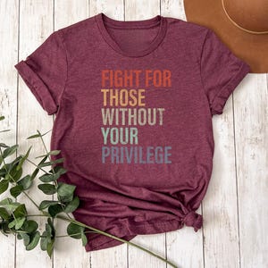 May include: Burgundy t-shirt with the text "FIGHT FOR THOSE WITHOUT YOUR PRIVILEGE" in orange, yellow, gray, and blue. The shirt is knotted at the bottom. Green leafy branches are in the lower left corner, and a brown hat is in the upper right corner.