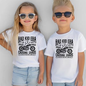 May include: Two children wearing white t-shirts with a black and white graphic of a motorcycle and the text "Rad Kid Era Causing Chaos Life in the Fast Lane".