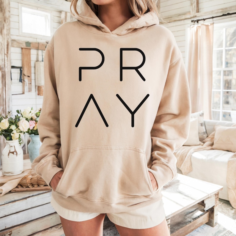 May include: A beige hoodie with the word "PRAY" in black block letters. The hoodie has a hood, a front pocket, and long sleeves. The model is wearing white shorts. The background is a light-colored room with a wooden table.