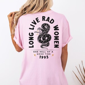 Peut inclure: T-shirt rose clair avec l'inscription "LONG LIVE RAD WOMEN" et un motif de serpent noir. Le texte "ONE HELL OF A GOOD TIME 1993" est &eacute;galement pr&eacute;sent. Le t-shirt a des manches courtes et une coupe ample.