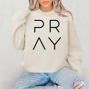 May include: Cream-colored sweatshirt with the word "PRAY" printed in black block letters. The sweatshirt has a crew neck and long sleeves. The model is wearing blue jeans.