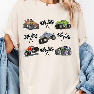 Pode incluir: Camiseta creme com seis desenhos coloridos de monster trucks com bandeiras quadriculadas. Os caminh&otilde;es s&atilde;o em v&aacute;rias cores, incluindo laranja, verde, vermelho e roxo, com gr&aacute;ficos detalhados. Um design divertido e l&uacute;dico.