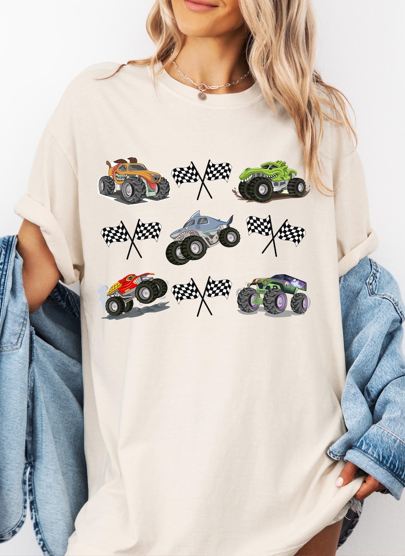Pode incluir: Camiseta creme com seis desenhos coloridos de monster trucks com bandeiras quadriculadas. Os caminh&otilde;es s&atilde;o em v&aacute;rias cores, incluindo laranja, verde, vermelho e roxo, com gr&aacute;ficos detalhados. Um design divertido e l&uacute;dico.