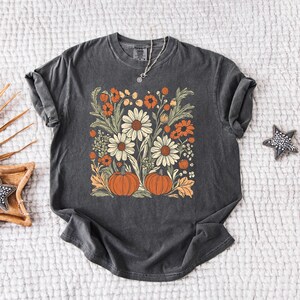 May include: Dark grey t-shirt featuring a floral design with daisies, orange flowers, and pumpkins. The design is centered on the shirt and has a vintage, slightly faded look. A silver necklace is visible at the neckline.