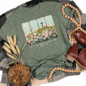 May include: An olive green t-shirt featuring a graphic of a cross on a hill with flowers and the text "HE IS NOT HERE. HE HAS RISEN. Matthew 28:6." The shirt is styled with brown shoes, wooden beads, and a decorative woven basket.