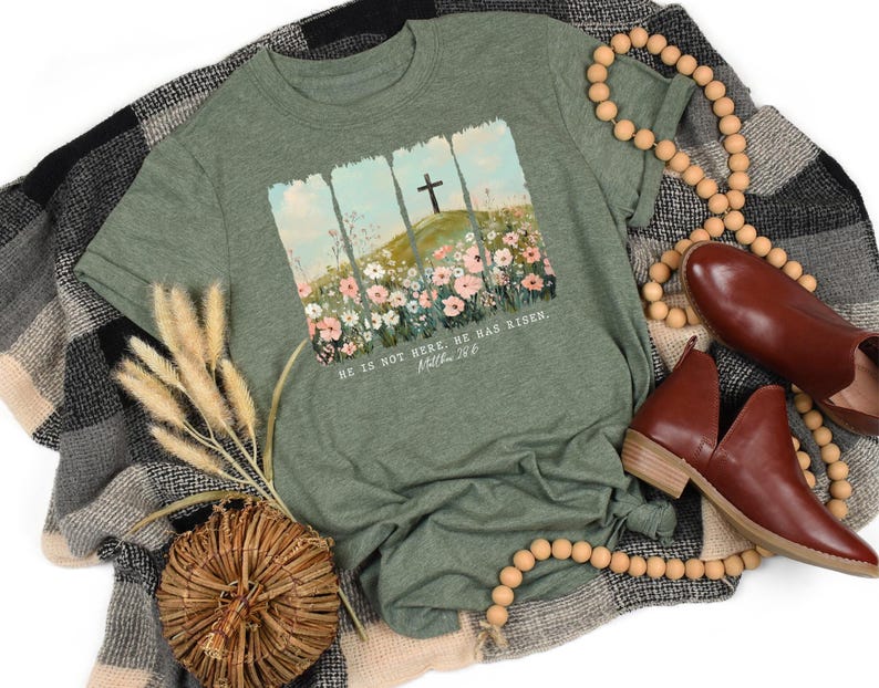 May include: An olive green t-shirt featuring a graphic of a cross on a hill with flowers and the text "HE IS NOT HERE. HE HAS RISEN. Matthew 28:6." The shirt is styled with brown shoes, wooden beads, and a decorative woven basket.