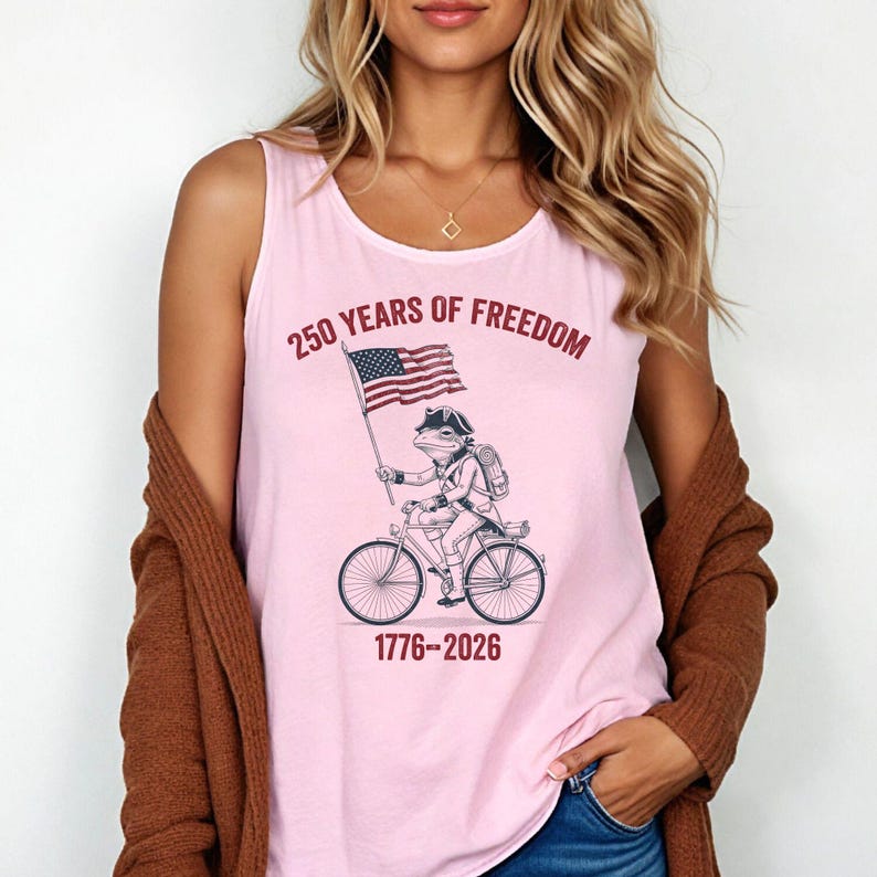 May include: Pink tank top with the text "250 YEARS OF FREEDOM" and the years "1776-2026". The graphic shows a frog riding a bicycle and holding an American flag.