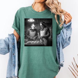 Puede incluir: Camiseta verde con un gr&aacute;fico en blanco y negro de dos figuras con delantales, una de ellas con una m&aacute;scara de hockey, preparando comida en una cocina. La camiseta tiene un corte holgado y mangas enrolladas.