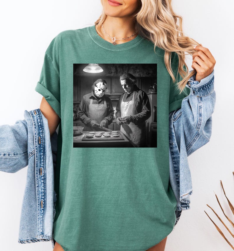 Puede incluir: Camiseta verde con un gr&aacute;fico en blanco y negro de dos figuras con delantales, una de ellas con una m&aacute;scara de hockey, preparando comida en una cocina. La camiseta tiene un corte holgado y mangas enrolladas.