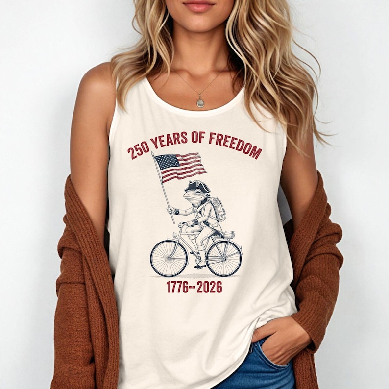 May include: Cream-colored tank top featuring a graphic of a frog riding a bicycle and holding an American flag. The text "250 YEARS OF FREEDOM" and "1776-2026" are printed above and below the graphic, respectively.