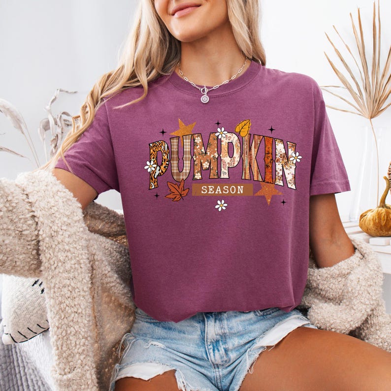 Pu&ograve; includere: Una maglietta bordeaux con la scritta "PUMPKIN SEASON" in un design a tema autunnale. Le lettere sono decorate con stampa leopardata, quadri e motivi floreali. La maglietta ha maniche corte e girocollo.