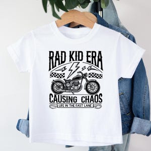 May include: White t-shirt with a black graphic of a motorcycle and the text "Rad Kid Era Causing Chaos Life in the Fast Lane"