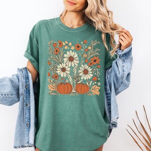 May include: A green t-shirt featuring a floral design with white daisies, orange flowers, and two orange pumpkins. The design is set against a square background. The shirt has rolled-up sleeves.