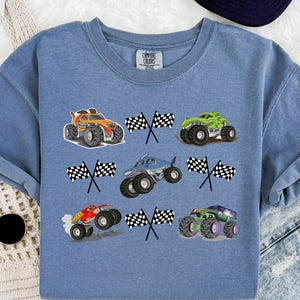 Pode incluir: Camisola azul Comfort Colors com seis gr&aacute;ficos coloridos de monster trucks e bandeiras quadriculadas. Os caminh&otilde;es s&atilde;o laranja, verde, azul, vermelho e roxo. Gola redonda e mangas compridas.
