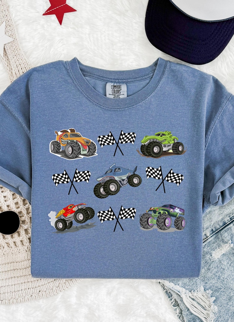 Pode incluir: Camisola azul Comfort Colors com seis gr&aacute;ficos coloridos de monster trucks e bandeiras quadriculadas. Os caminh&otilde;es s&atilde;o laranja, verde, azul, vermelho e roxo. Gola redonda e mangas compridas.