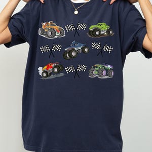 Pode incluir: Camiseta azul marinho com gr&aacute;ficos coloridos de monster trucks e bandeiras quadriculadas. Os monster trucks s&atilde;o nas cores laranja, verde, azul, vermelho e roxo. A camiseta tem gola redonda e mangas curtas.