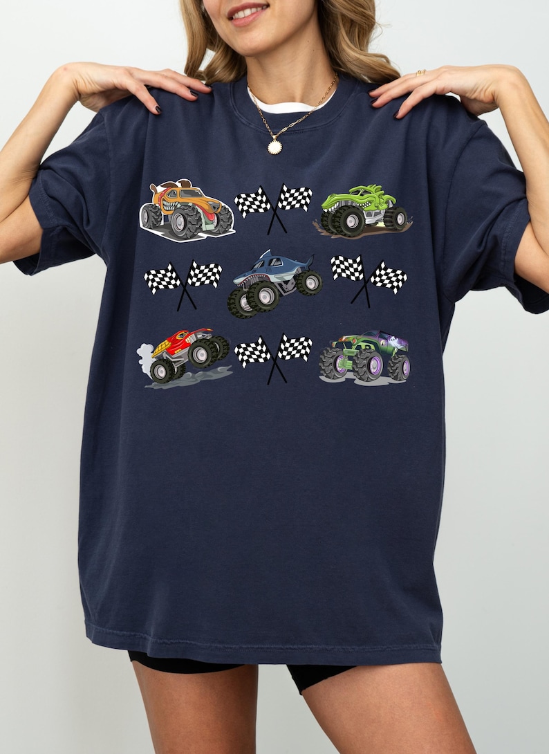 Pode incluir: Camiseta azul marinho com gr&aacute;ficos coloridos de monster trucks e bandeiras quadriculadas. Os monster trucks s&atilde;o nas cores laranja, verde, azul, vermelho e roxo. A camiseta tem gola redonda e mangas curtas.
