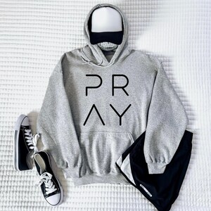 May include: A gray hoodie with the word "PRAY" in black block letters, a white and black baseball cap, black shorts, and black and white sneakers are arranged on a white textured surface. The hoodie has a front pocket.