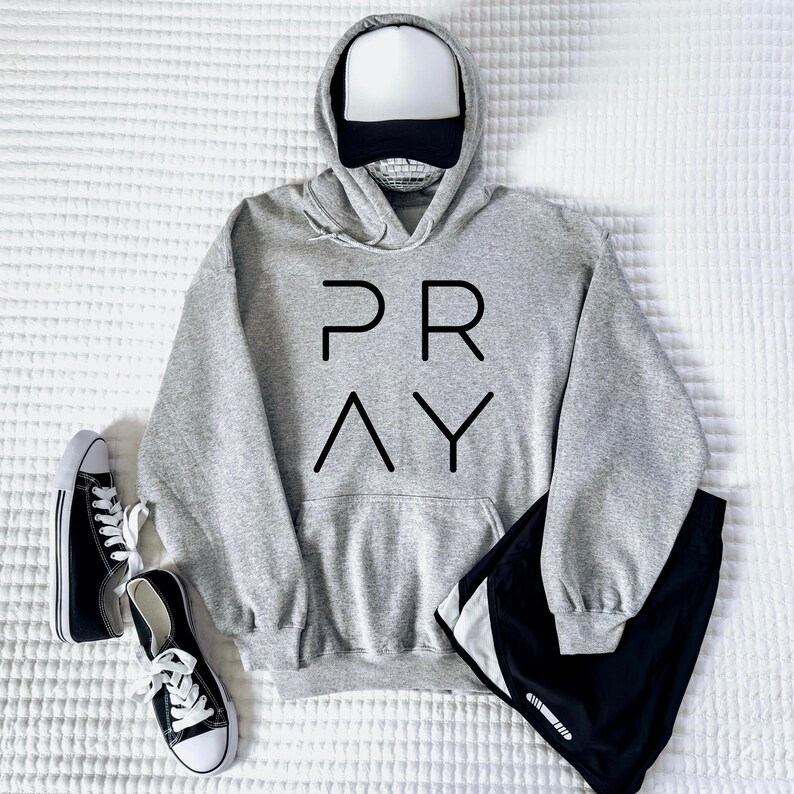 May include: A gray hoodie with the word "PRAY" in black block letters, a white and black baseball cap, black shorts, and black and white sneakers are arranged on a white textured surface. The hoodie has a front pocket.