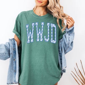 May include: A green t-shirt with the letters "WWJD" in a blue floral design. The shirt has a relaxed fit and short sleeves. A denim jacket is draped over the shoulders.