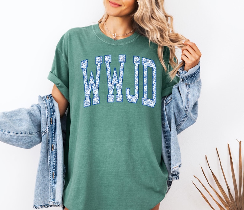 May include: A green t-shirt with the letters "WWJD" in a blue floral design. The shirt has a relaxed fit and short sleeves. A denim jacket is draped over the shoulders.