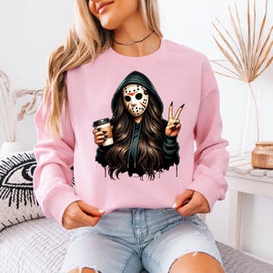 May include: A pale pink sweatshirt with a graphic of a person in a hockey mask and black hoodie, holding a coffee cup and making a peace sign. The graphic has long dark hair and a black and white mask.