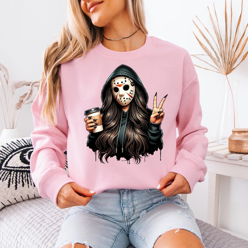 May include: A pale pink sweatshirt with a graphic of a person in a hockey mask and black hoodie, holding a coffee cup and making a peace sign. The graphic has long dark hair and a black and white mask.