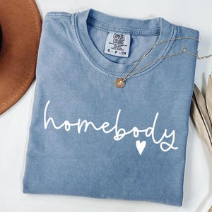 May include: A light blue t-shirt with the text "homebody" in white script with a small white heart.