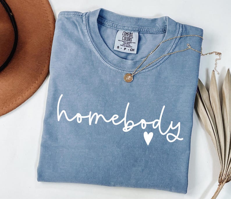 May include: A light blue t-shirt with the text "homebody" in white script with a small white heart.