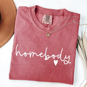 May include: A red, vintage-style t-shirt with the word "homebody" written in white cursive lettering with a small white heart underneath.