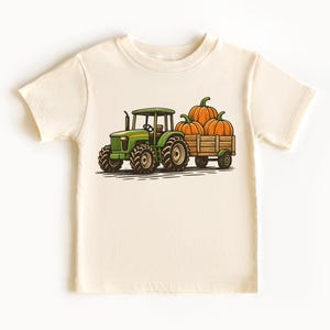 May include: Cream-colored t-shirt featuring a green tractor pulling a wooden wagon filled with three orange pumpkins. The tractor has large black tires and a detailed design. A fall-themed graphic tee for kids.