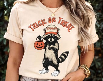 May include: Beige t-shirt with the words "Trick or Treat" in orange lettering. A cartoon raccoon wearing a hat that says "Spooky" and holding a jack-o'-lantern bucket is featured on the front. The raccoon is wearing white clogs.