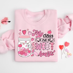 Pu&ograve; includere: Felpa rosa con una grafica di un barattolo pieno di cuori e il testo "My class is full of sweet hearts".